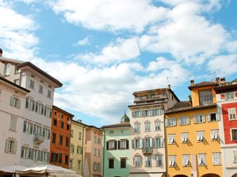 Reihe bunter historischer Gebäude am Domplatz in Trient mit grünen, gelben, roten und weißen Fassaden unter blauem Himmel.