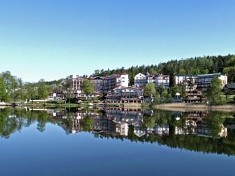 Blick über einen ruhigen See, der das Hotel Solar und umliegende Gebäude an einem bewaldeten Hang in Masuren, Polen, unter blauem Himmel spiegelt.