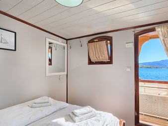 Helle Doppelkabine auf MY Magellan mit weißer Bettwäsche, Holzverkleidung, Bullauge und Tür zum Deck mit Blick auf die blaue Adria.