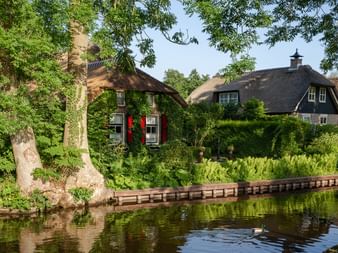 Traditionelle niederländische Reetdachhäuser entlang eines Kanals in Giethoorn. Bäume rahmen die Szene ein, eine Ente schwimmt im Wasser.
