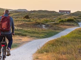Radfahrer mit rotem Rucksack schiebt Fahrrad auf gewundenem Weg durch grasbewachsene Dünen auf Terschelling bei Sonnenuntergang.