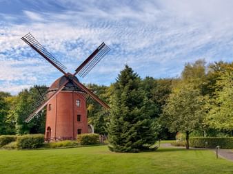 Rote Backsteinwindmühle mit vier Flügeln in einem grünen Park in Rastede, umgeben von Bäumen und Rasen unter blauem Himmel mit weißen Wolken.
