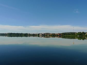 Panoramablick auf einen ruhigen See in Masuren, Polen, mit bunten Häusern und Bäumen, die sich im stillen Wasser unter blauem Himmel spiegeln.