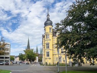 Gelbes historisches Turmgebäude mit dunkler Kuppel in Oldenburg. Kirchtürme im Hintergrund, große Bäume rahmen die Szene unter blauem Himmel.