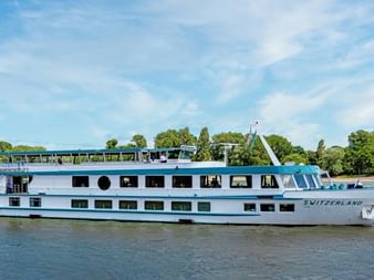 Weißes Flusskreuzfahrtschiff MS Switzerland mit blauem Zierstreifen auf ruhigem Wasser. Bäume und blauer Himmel im Hintergrund.