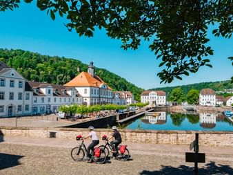 Zwei Radfahrer mit Rädern ruhen an Steinmauer mit Blick auf Hafen in Bad Karlshafen. Historische weiße Barockgebäude säumen das Ufer unter blauem Himmel.