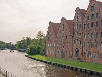 Reihe historischer roter Backsteingebäude mit Treppengiebeln entlang eines Wasserwegs in Lübeck. Grüne Rasenfläche und Holzsteg am Wasser.