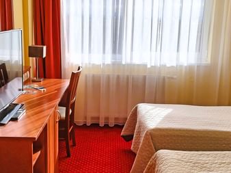 Helles Hotelzimmer mit zwei Einzelbetten, Holzschreibtisch, Stuhl, TV, rotem Teppich und weißen Gardinen mit orangen Vorhängen im Hotel Ludmila in Mělník.