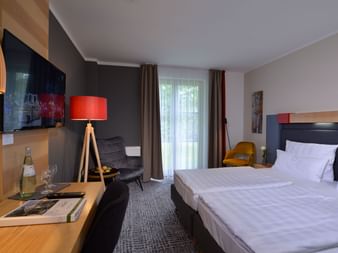 Zeitgenössisches Hotelzimmer mit weißem Bett, dunkler Akzentwand, Holzschreibtisch, Wandfernseher, Stativlampe und Sessel am Fenster.