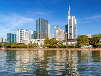 Frankfurter Skyline mit modernen Wolkenkratzern spiegelt sich im Main. Grüne Bäume säumen das Flussufer unter blauem Himmel mit Wolken.