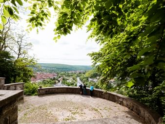 Steinerner Aussichtspunkt mit Besuchern, die durch grünes Blätterdach auf Hann. Münden blicken. Runde Steinterrasse mit niedrigen Mauern.