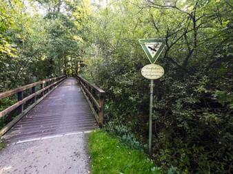 Holzbrücke auf einem Radweg bei Rastede, umgeben von dichtem grünem Wald. Ein Vorfahrtsschild und ovales Schild sind rechts sichtbar.