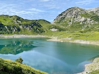 Formarinsee, ein türkisfarbener Alpensee umgeben von grünen Wiesen und felsigen Berggipfeln unter blauem Himmel entlang des Lechradwegs in Österreich.