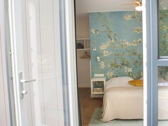 Helles Hotelzimmer mit Bett, Blumentapete in Blau- und Goldtönen, weißem Nachttisch und Glastüreingang im Hotel Jaans, Gaasterland.