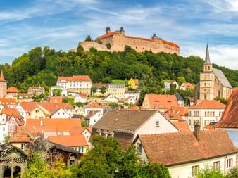 Panoramablick auf Kulmbach mit historischer Burg auf dem Hügel über der Stadt mit roten Ziegeldächern, Kirchturm und grünen Wäldern.