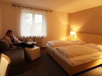 Hotelzimmer im Hotel Central Zeven mit Doppelbett, grauem Sofa, Leselampe und Fenster mit weißen Vorhängen. Gast entspannt im Sitzbereich.