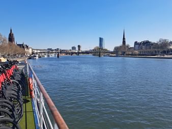 Reihe von Fahrrädern mit roten Taschen auf dem Deck der MS Casanova entlang eines Flusses. Stadtsilhouette mit Kirchtürmen und Brücke im Hintergrund.