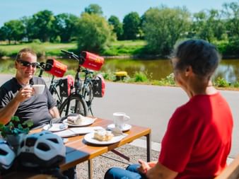 Zwei Radfahrer genießen Kaffee an Außentisch nahe der Weser in Höxter. Fahrräder mit roten Taschen dahinter, grüne Landschaft sichtbar.
