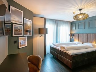 Modernes Hotelzimmer mit türkisfarbenen Wänden, beigem Polsterbett, dekorativer Deckenleuchte, gerahmten Fotos, Stehlampe und braunem Sessel.