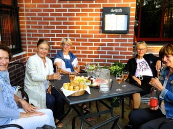 Fünf Frauen sitzen an einem Tisch im Freien mit Getränken und Snacks vor einer roten Backsteinmauer und genießen einen entspannten Moment.