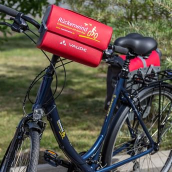 Blaues Tourenfahrrad mit schwarzem Gepäckträger und roter Rückenwind-Lenkertasche mit Vaude-Logo, auf Gras geparkt mit Bäumen im Hintergrund.