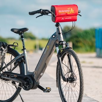 Graues Fahrrad mit roten Rückenwind-Taschen am Lenker und Gepäckträger, auf Holzsteg am Strand mit Umkleidekabinen im Hintergrund.