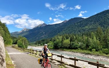 Radfahrer mit Helm und Packtaschen auf rotem Fahrrad neben Holzgeländer entlang Fluss mit bewaldeten Bergen und blauem Himmel.