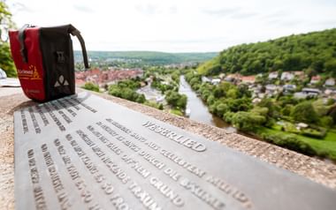 Metalltafel mit deutschem Text an Aussichtspunkt über Hann. Münden Stadt und Flusstal, mit roten Fahrradtaschen sichtbar.