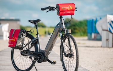 Gray bicycle with red Rückenwind panniers on handlebar and rear rack, parked on wooden boardwalk near beach with changing cabins in background.
