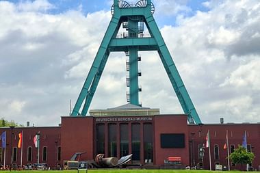 Ikonischer türkisfarbener Förderturm überragt das rote Backsteingebäude des Deutschen Bergbau-Museums in Bochum. Grüne Wiese im Vordergrund.