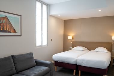 Modernes Hotelzimmer in Toulouse mit zwei Einzelbetten, grauem Sofa, Wandlampen und gerahmtem Kunstwerk mit historischem Gebäude.