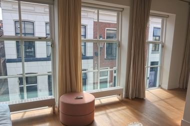 Helles Hotelzimmer in Kampen mit drei großen Fenstern mit Blick auf einen Kanal und historische Gebäude. Heller Holzboden und beige Vorhänge.