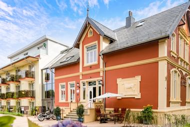 Orangefarbenes historisches Gebäude mit cremefarbenen Verzierungen und grauem Dach, Parkhotel Jever. Modernes weißes Hotelgebäude dahinter.