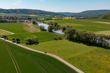 Aerial view of cyclists on a dirt path through green fields in the Weserbergland. River Weser winds through the valley with hills and villages.