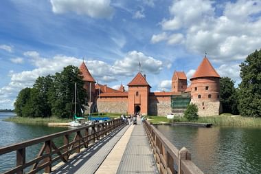 Holzsteg zur Trakai Inselburg mit roten Backsteintürmen und Mauern, umgeben von Seewasser und grünen Bäumen unter blauem Himmel.