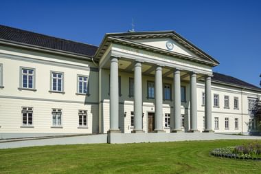 Weißes klassizistisches Schlossgebäude mit sechs hohen Säulen, die einen dreieckigen Giebel stützen. Das zweistöckige Gebäude hat symmetrische Fenster und steht auf gepflegtem grünen Rasen unter blauem Himmel.