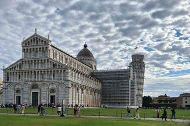 Dom von Pisa mit weißer Marmorfassade und der berühmte Schiefe Turm von Pisa auf grüner Wiese unter dramatischem Wolkenhimmel mit Touristen.
