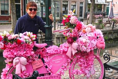 Lächelnde Frau mit Sonnenbrille neben einem rosa Fahrrad mit rosa Blumen an einem Kanal in Edam, Niederlande.