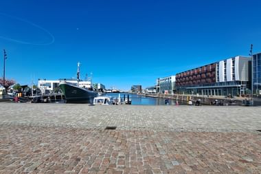 Kopfsteinpflaster-Hafenplatz mit historischem Schiff, rot-weißem Leuchtturm und modernen Ufergebäuden unter blauem Himmel in Bremerhaven.