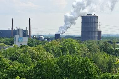 Luftaufnahme der Kokerei Prosper mit dampfendem Kühlturm, Schornsteinen und Gebäuden, umgeben von dichtem Grün.