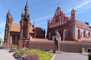Roter gotischer St.-Anna-Kirchenkomplex in Vilnius mit verzierten Türmen, angrenzender Bernhardinerkirche und Statue im Vordergrund.