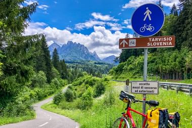Rotes Tourenrad mit gelben Packtaschen neben Wegweiser nach Tarvisio Slowenien auf Bergradweg mit Alpengipfeln im Hintergrund.