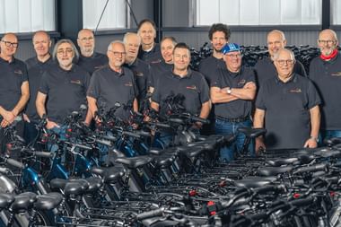 Gruppe von dreizehn Stationsleitern in dunklen Poloshirts stehen hinter Reihen schwarzer und blauer Mietfahrräder in einer Lagerhalle.