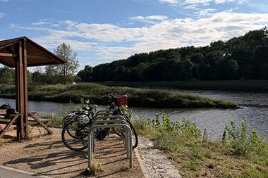 Fahrrad-Rastplatz bei Melnik mit Holzunterstand und Fahrradständer am Fluss. Zwei Fahrräder geparkt, bewaldete Hügel im Hintergrund unter blauem Himmel.