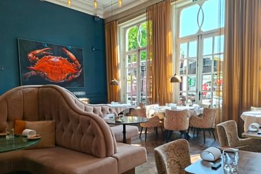 Eleganter Frühstücksraum mit türkisfarbenen Wänden, beigen Polstersitzen und großen Fenstern mit goldenen Vorhängen. Ein rotes Krabben-Kunstwerk hängt an der Wand.