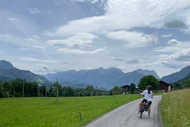 Radfahrer auf asphaltierter Landstraße durch grüne Wiesen mit Alpengebirgskette im Hintergrund unter bewölktem Himmel.