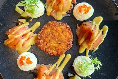 Gourmet-Vorspeise auf schwarzem Schieferplatte mit knusprigem paniertem Plätzchen in der Mitte, umgeben von Räucherlachs und Frischkäse mit Kaviar.
