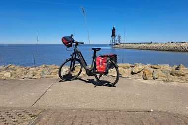 Reiserad mit roten Packtaschen auf Hafenpromenade in Dorum-Neufeld. Steinmole und Leuchtturm über ruhigem blauen Wasser sichtbar.