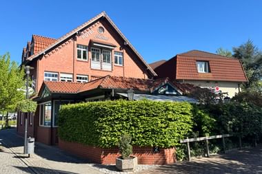 Rotes Backsteingebäude Hotel Central in Zeven mit orangefarbenem Ziegeldach und grüner Hecke. Klarer blauer Himmel und weißes Nebengebäude.