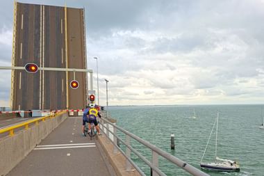 Radfahrer wartet an geöffneter Klappbrücke der Zeelandbrücke bei roten Ampeln. Segelboot passiert die Wasserstraße darunter unter bewölktem Himmel.